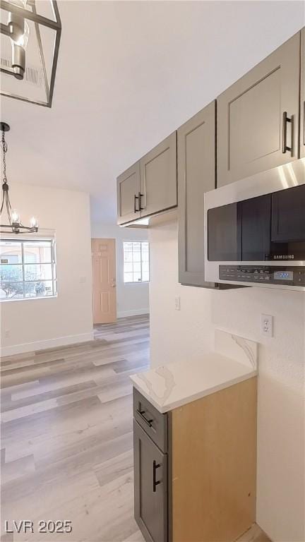 kitchen with light wood finished floors, gray cabinets, stainless steel microwave, hanging light fixtures, and an inviting chandelier