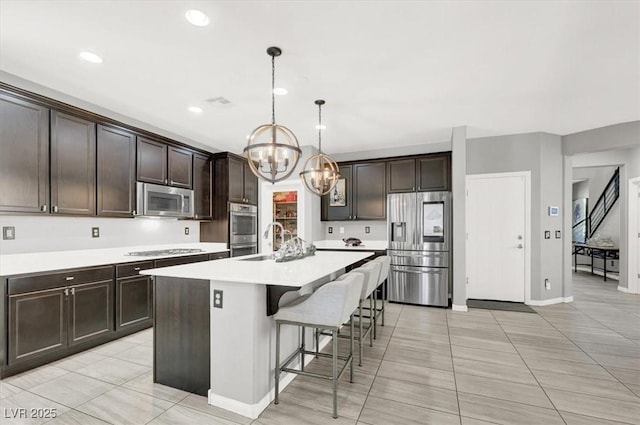 kitchen with dark brown cabinetry, light countertops, appliances with stainless steel finishes, an island with sink, and a kitchen bar