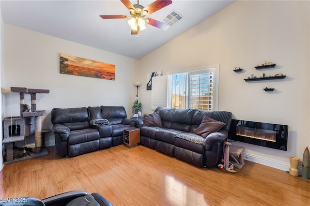 living room with visible vents, ceiling fan, light wood-type flooring, vaulted ceiling, and a glass covered fireplace