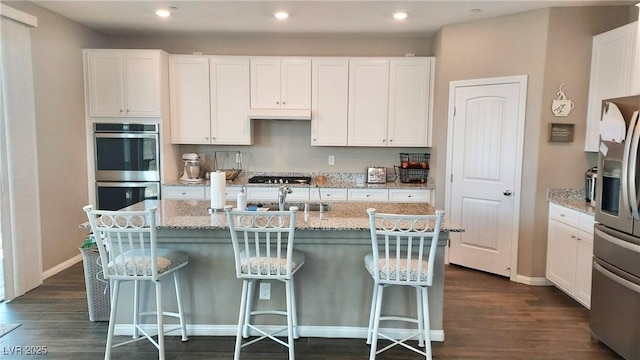 kitchen featuring appliances with stainless steel finishes, dark wood-style flooring, a kitchen island with sink, and light stone counters