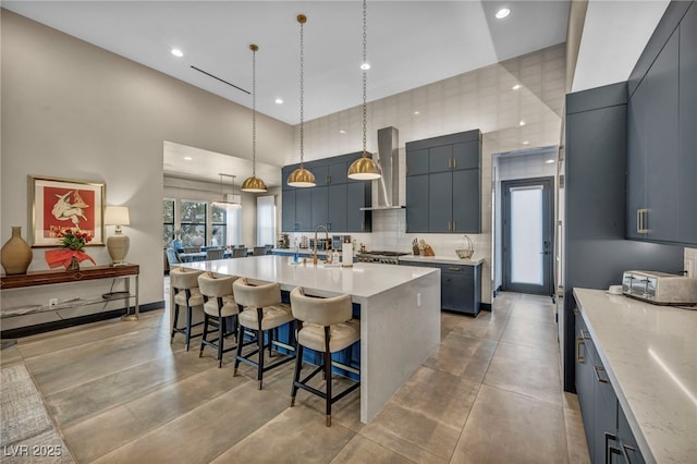 kitchen with hanging light fixtures, backsplash, wall chimney range hood, an island with sink, and a kitchen bar