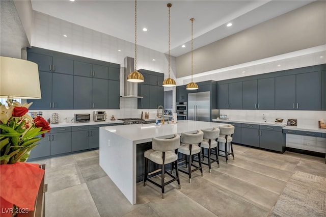 kitchen with stainless steel appliances, light countertops, a towering ceiling, wall chimney range hood, and a kitchen bar