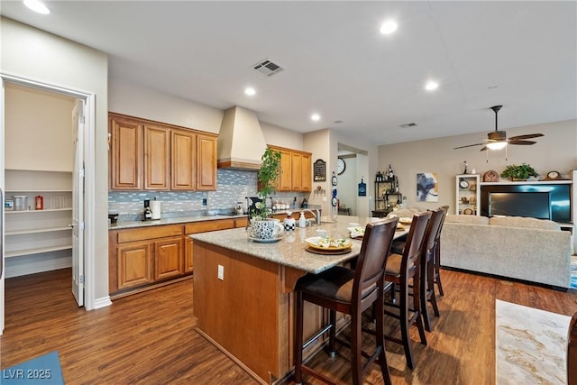 kitchen featuring custom exhaust hood, a center island with sink, visible vents, and dark wood-style flooring