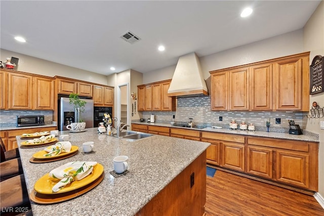 kitchen with wood finished floors, a sink, visible vents, black appliances, and custom range hood