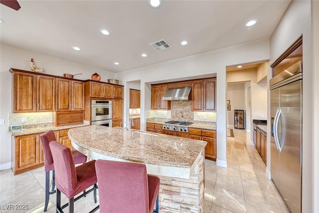 kitchen with brown cabinets, a center island with sink, stainless steel appliances, a sink, and wall chimney exhaust hood