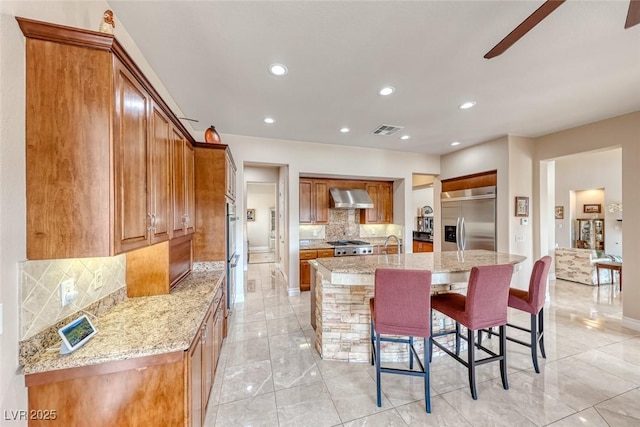 kitchen featuring a breakfast bar, appliances with stainless steel finishes, wall chimney range hood, brown cabinetry, and a large island with sink