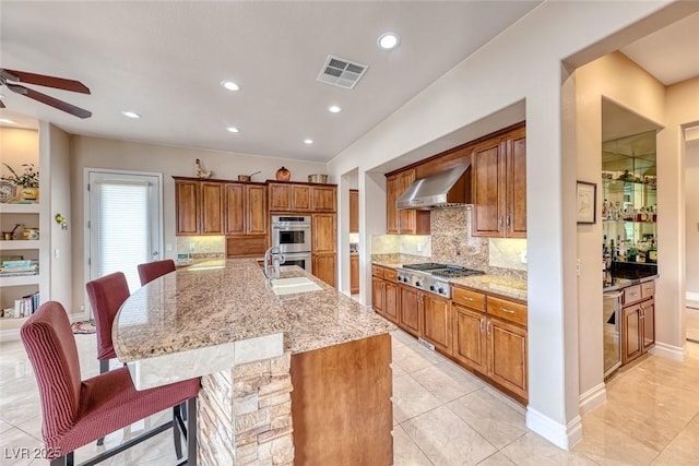 kitchen featuring visible vents, wall chimney range hood, appliances with stainless steel finishes, backsplash, and brown cabinetry