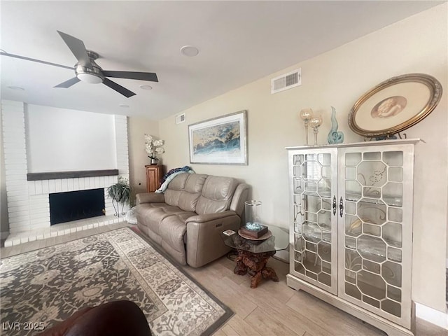 living room with a ceiling fan, a brick fireplace, visible vents, and wood finished floors