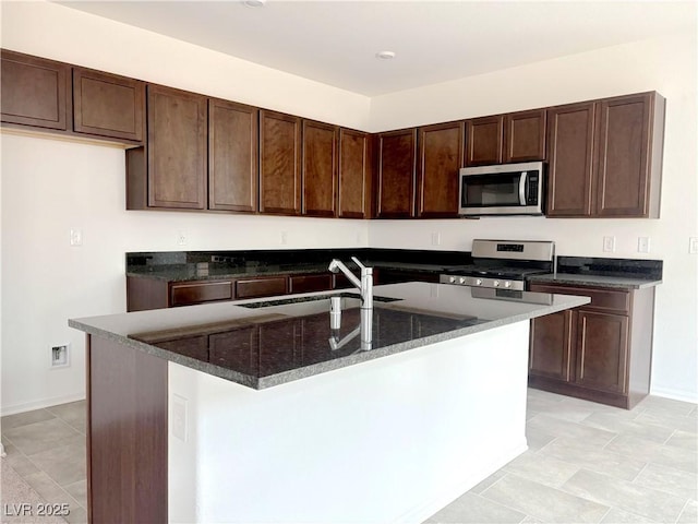 kitchen featuring appliances with stainless steel finishes, a sink, dark brown cabinetry, dark stone countertops, and an island with sink