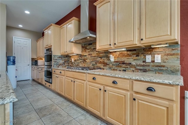 kitchen with stainless steel appliances, wall chimney exhaust hood, light brown cabinets, light stone counters, and backsplash