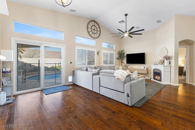 living area with a warm lit fireplace, high vaulted ceiling, ceiling fan, dark wood-type flooring, and visible vents