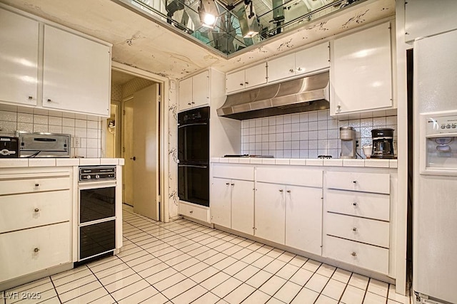 kitchen featuring white refrigerator with ice dispenser, tile countertops, dobule oven black, white cabinets, and under cabinet range hood