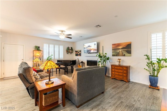 living room featuring light wood-type flooring, a glass covered fireplace, visible vents, and a ceiling fan