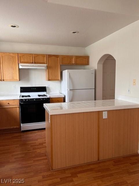 kitchen with tile counters, dark wood-type flooring, freestanding refrigerator, gas range, and under cabinet range hood