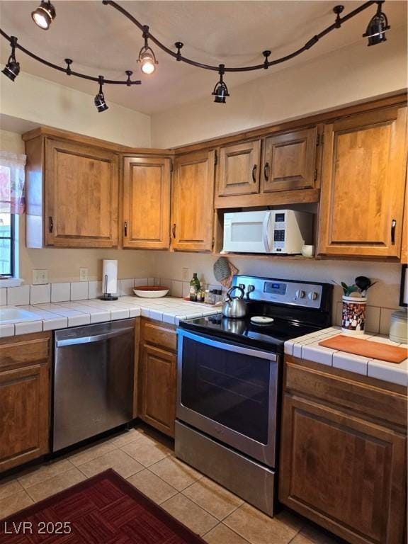 kitchen with tile countertops, appliances with stainless steel finishes, brown cabinetry, and light tile patterned floors