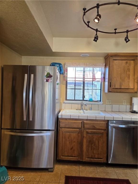 kitchen featuring light tile patterned floors, tile counters, appliances with stainless steel finishes, brown cabinets, and a sink