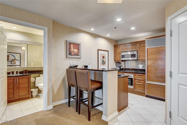 kitchen featuring dark countertops, brown cabinets, appliances with stainless steel finishes, a kitchen breakfast bar, and light tile patterned flooring