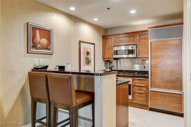 kitchen with brown cabinetry, dark countertops, paneled built in fridge, a kitchen bar, and stainless steel microwave