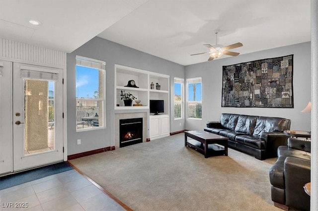 carpeted living area featuring a warm lit fireplace, baseboards, ceiling fan, tile patterned flooring, and built in shelves