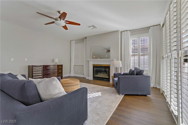 living area featuring visible vents, a glass covered fireplace, ceiling fan, wood finished floors, and baseboards