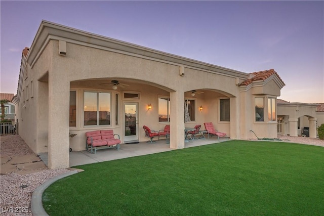 back of house at dusk with ceiling fan, a patio area, a lawn, and stucco siding