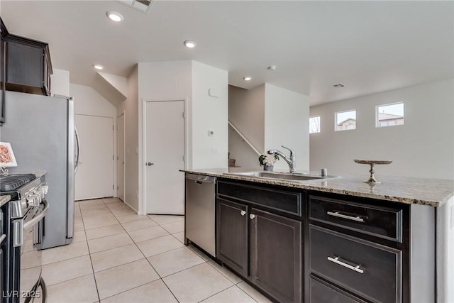 kitchen with light tile patterned floors, a kitchen island with sink, a sink, appliances with stainless steel finishes, and light stone countertops