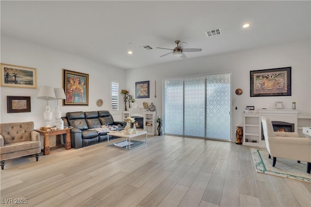 living area featuring light wood-type flooring, a glass covered fireplace, visible vents, and a ceiling fan