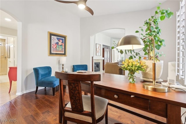 home office with arched walkways, dark wood-style flooring, a ceiling fan, baseboards, and a glass covered fireplace