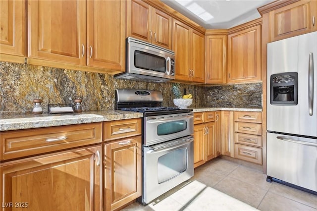 kitchen featuring decorative backsplash, brown cabinets, light stone countertops, stainless steel appliances, and light tile patterned flooring