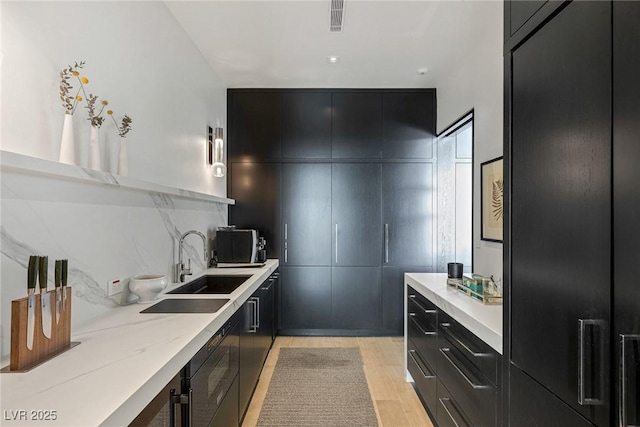 kitchen with dark cabinetry, light stone countertops, light wood finished floors, a sink, and decorative backsplash