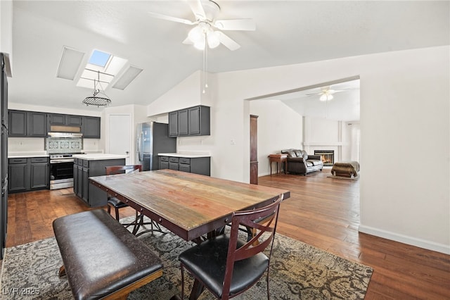 dining room with dark wood-style floors, vaulted ceiling with skylight, a ceiling fan, and a glass covered fireplace