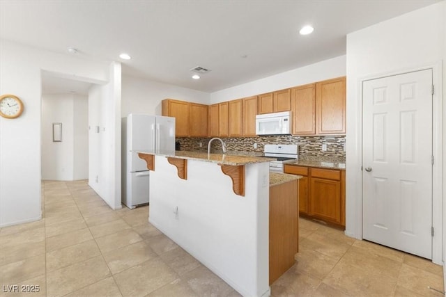 kitchen with light stone counters, a center island with sink, backsplash, white appliances, and a kitchen bar