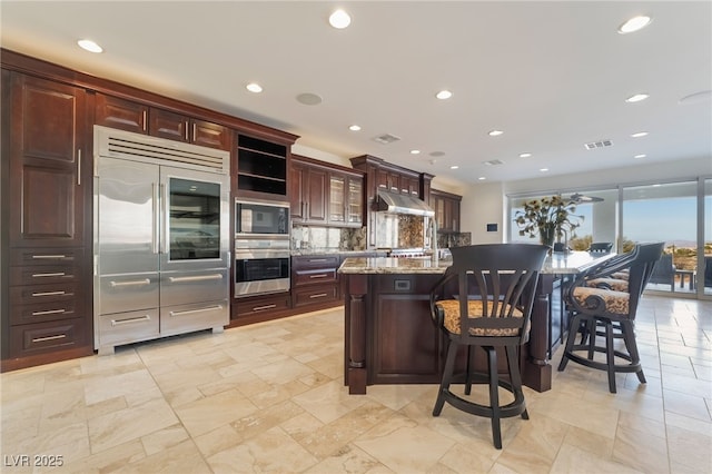 kitchen featuring backsplash, an island with sink, built in appliances, under cabinet range hood, and a kitchen breakfast bar