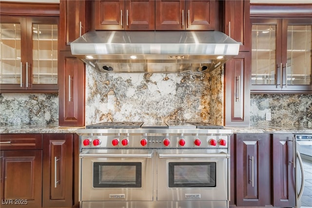 kitchen featuring reddish brown cabinets, light stone countertops, under cabinet range hood, double oven range, and backsplash