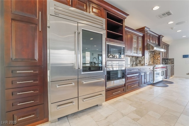 kitchen with visible vents, backsplash, glass insert cabinets, built in appliances, and under cabinet range hood