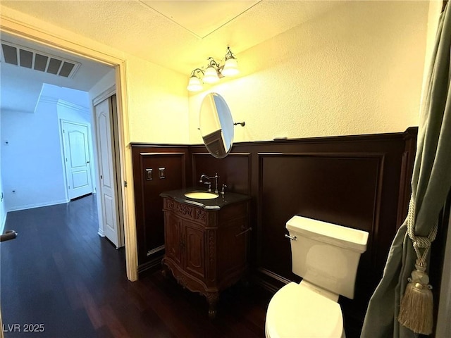 half bath with visible vents, toilet, wainscoting, vanity, and wood finished floors