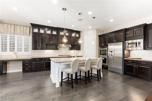 kitchen featuring a breakfast bar, dark wood finished floors, stainless steel appliances, a kitchen island with sink, and under cabinet range hood