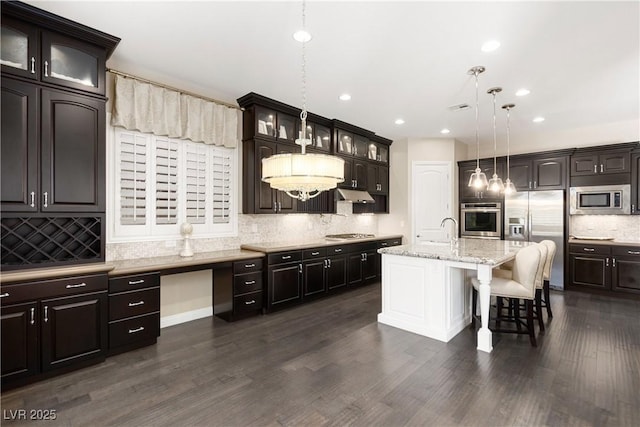 kitchen with dark wood-type flooring, a kitchen breakfast bar, appliances with stainless steel finishes, an island with sink, and glass insert cabinets