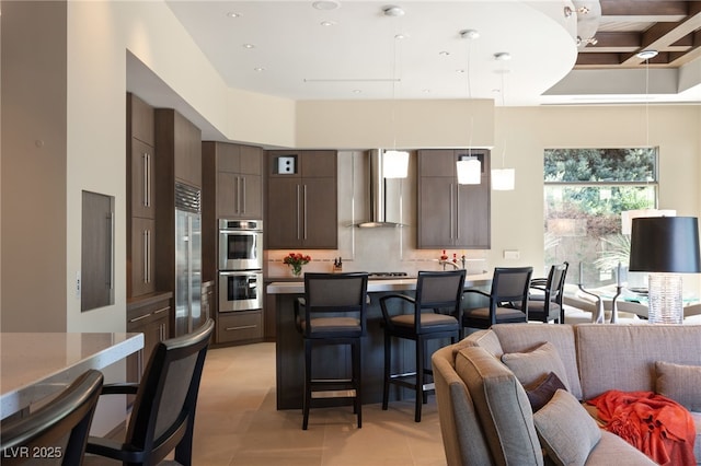 kitchen featuring a breakfast bar, dark brown cabinets, appliances with stainless steel finishes, backsplash, and wall chimney exhaust hood