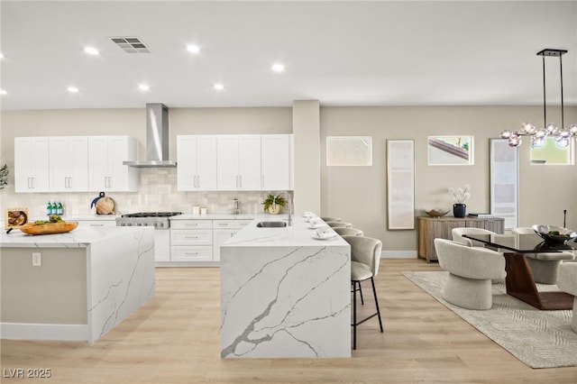 kitchen with a breakfast bar area, white cabinetry, visible vents, wall chimney range hood, and light stone countertops