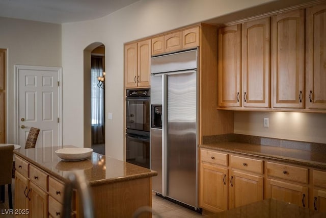 kitchen with light tile patterned floors, arched walkways, dark stone countertops, a center island, and built in fridge