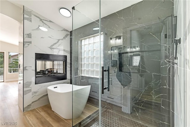 full bathroom with wood finished floors, tile walls, a multi sided fireplace, a freestanding bath, and a marble finish shower