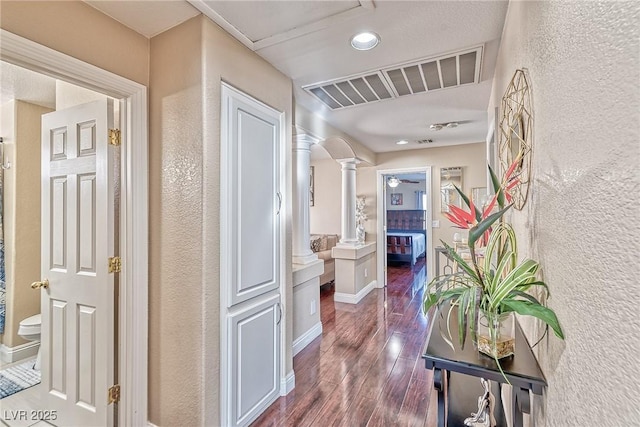 hall featuring a textured wall, dark wood-style flooring, visible vents, baseboards, and ornate columns