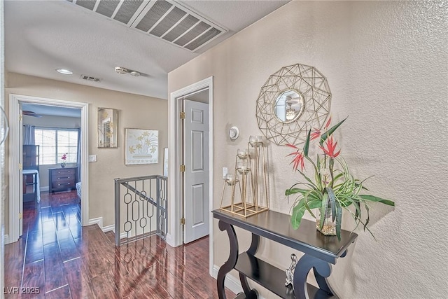 corridor with dark wood-style flooring, visible vents, a textured wall, an upstairs landing, and baseboards