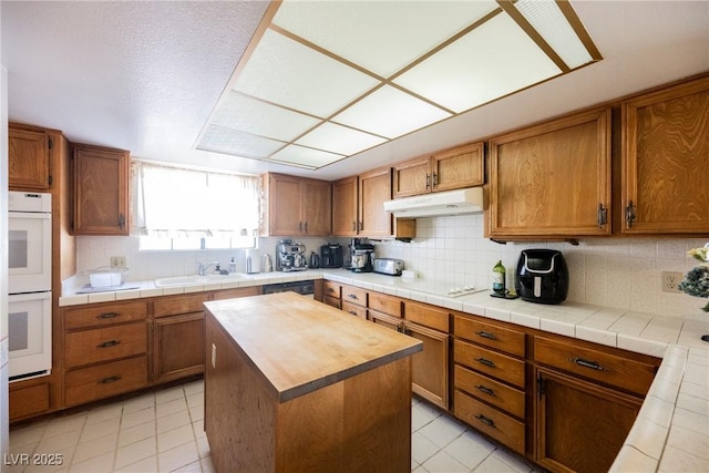 kitchen with white appliances, brown cabinetry, backsplash, under cabinet range hood, and a sink