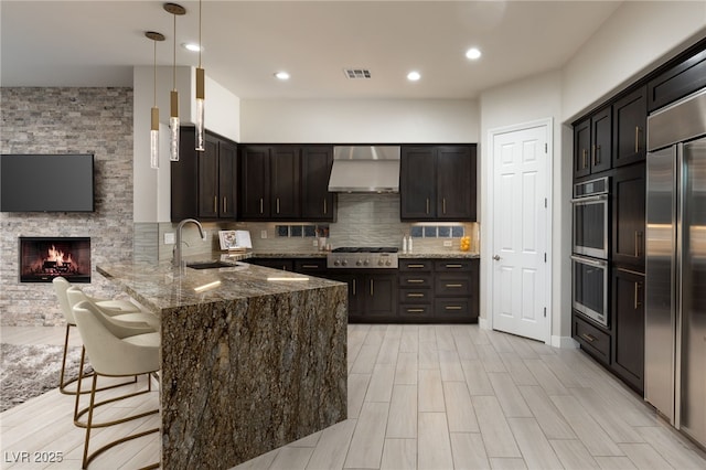 kitchen with a peninsula, a sink, appliances with stainless steel finishes, dark stone counters, and wall chimney exhaust hood