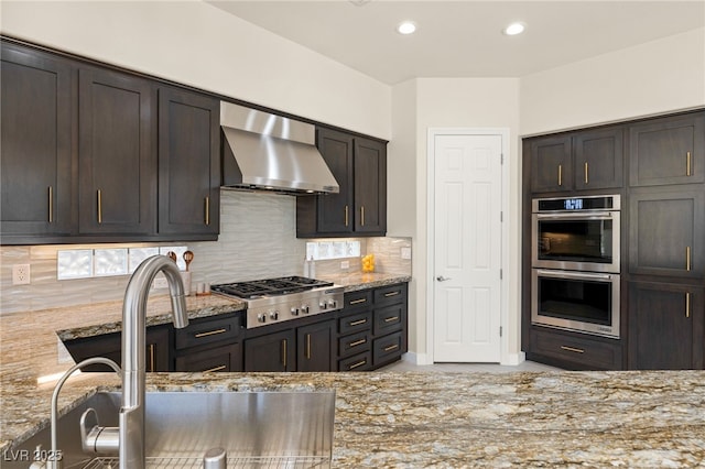 kitchen with decorative backsplash, wall chimney exhaust hood, appliances with stainless steel finishes, light stone countertops, and a sink