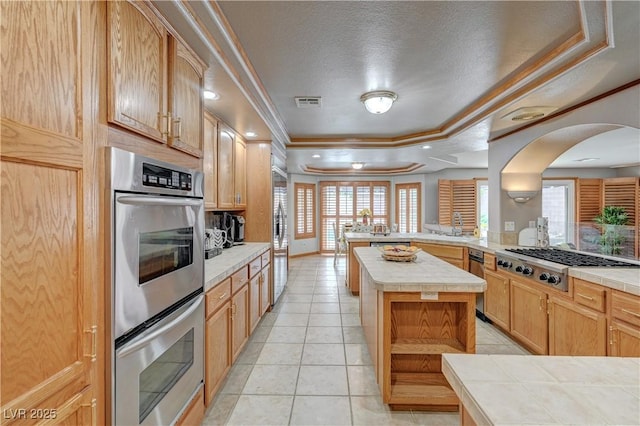 kitchen with tile counters, a raised ceiling, appliances with stainless steel finishes, a peninsula, and crown molding