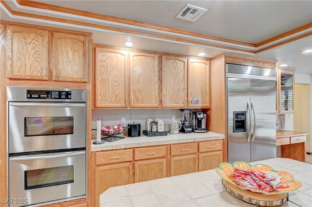 kitchen featuring visible vents, tile counters, ornamental molding, stainless steel appliances, and light brown cabinetry