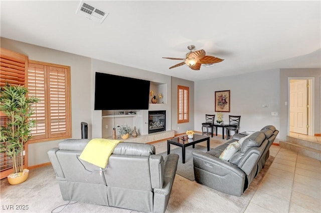 tiled living area featuring ceiling fan, visible vents, baseboards, and a glass covered fireplace